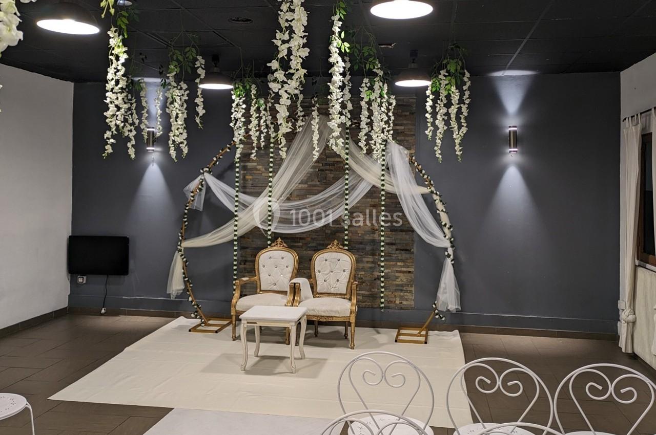 Deux chaises dorées avec une table blanche devant un décor floral suspendu dans une salle sombre.