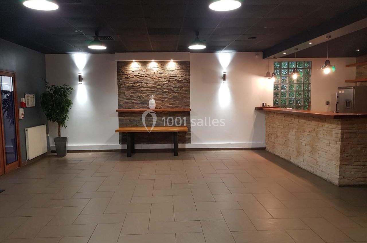 Salle vide avec sol carrelé, mur en pierre éclairé, banc en bois, plante en pot et comptoir en pierre à droite.