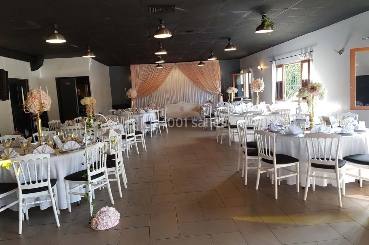 Salle de réception décorée avec des tables rondes, nappes blanches, chaises blanches et arrangements floraux.