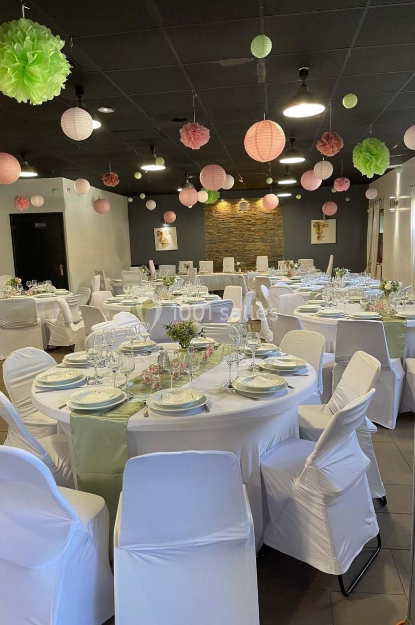 Salle décorée pour un événement, avec tables rondes dressées, nappes blanches et lanternes suspendues au plafond.