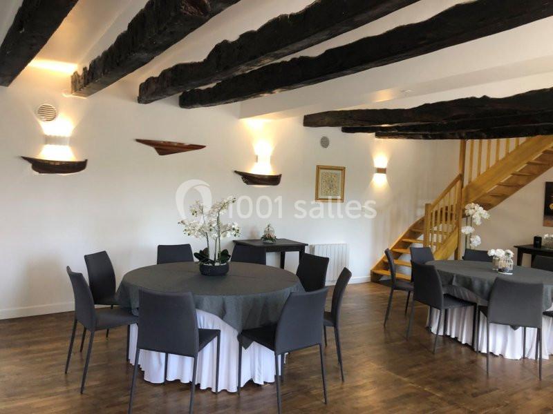 Salle lumineuse avec tables rondes dressées, chaises noires, poutres apparentes au plafond et escalier en bois.