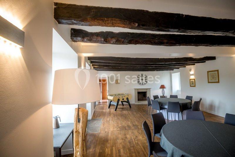 Salle lumineuse avec poutres apparentes, tables rondes dressées, baby-foot et cheminée au fond.