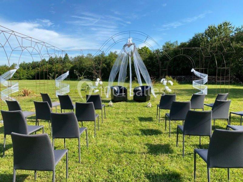 Chaises disposées en extérieur face à une arche décorée de voilages blancs et de fleurs, sur une pelouse verdoyante.