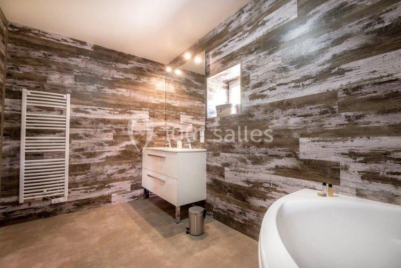 Salle de bain moderne avec murs effet bois, meuble blanc, miroir éclairé, radiateur et baignoire au premier plan.