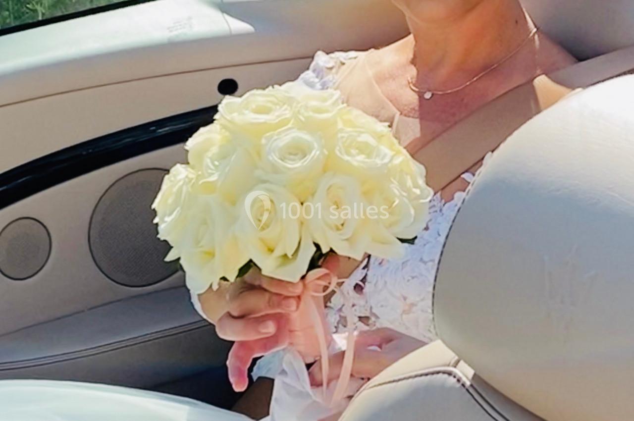Une femme en robe blanche tient un bouquet de roses blanches dans une voiture décapotable beige.