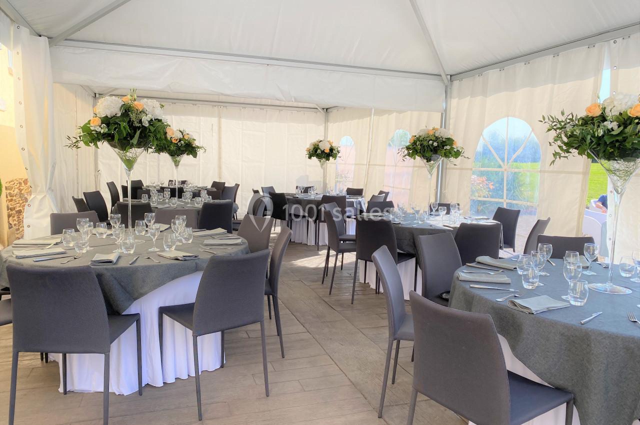 Salle de réception sous tente avec tables rondes dressées, nappes grises, chaises noires et décorations florales.