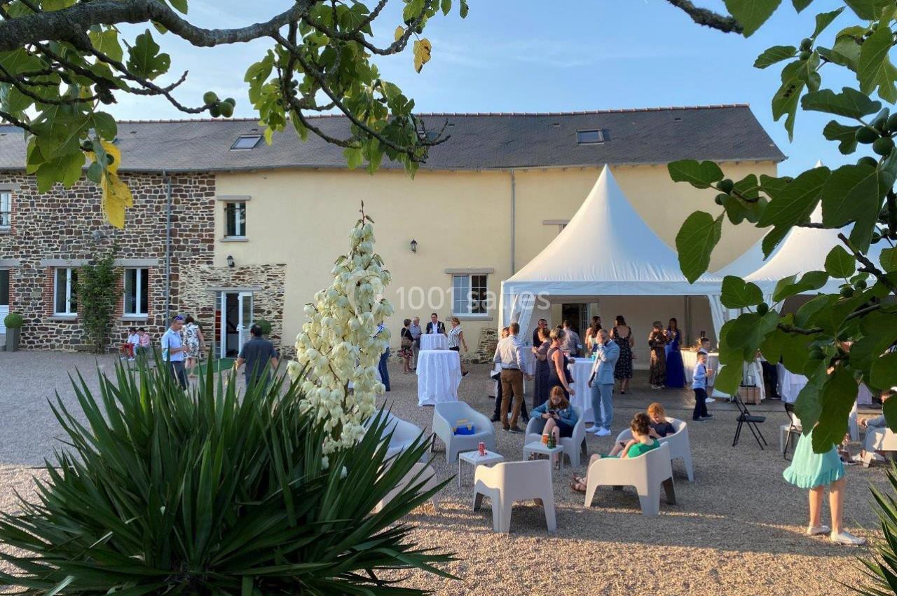 Personnes rassemblées dans une cour devant une maison en pierre et une tente blanche, par une journée ensoleillée.
