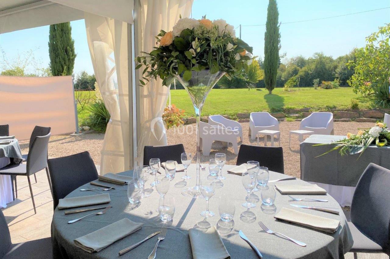 Table ronde élégamment dressée avec nappes grises, verres et couverts, sous une tente ouverte sur un jardin ensoleillé.
