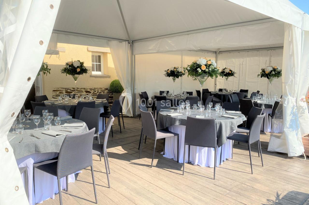 Tables rondes dressées avec nappes grises et décorations florales sous une tente blanche sur une terrasse ensoleillée.