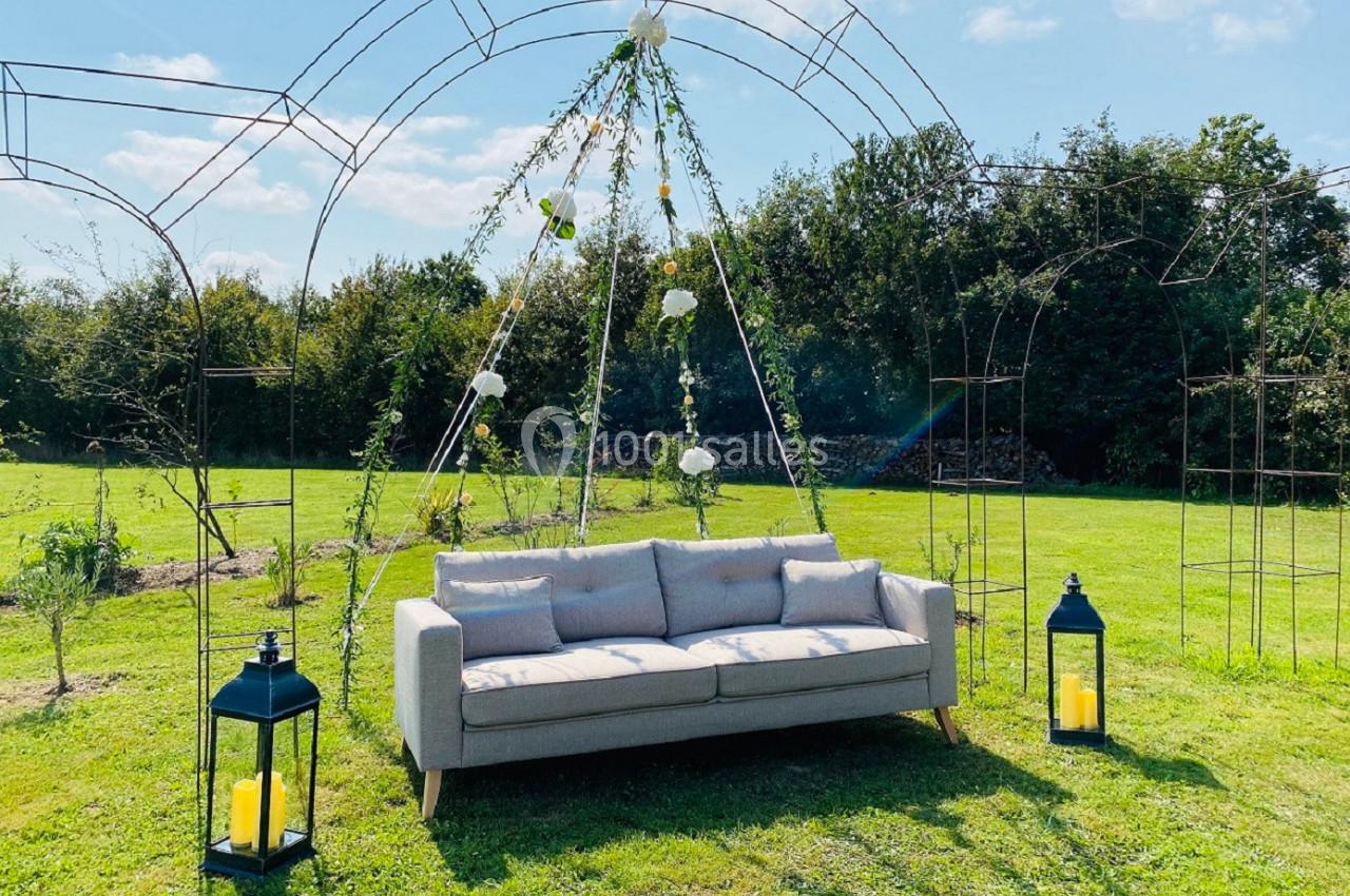 Canapé gris disposé sous une arche métallique décorée de fleurs et feuillages, dans un jardin ensoleillé.