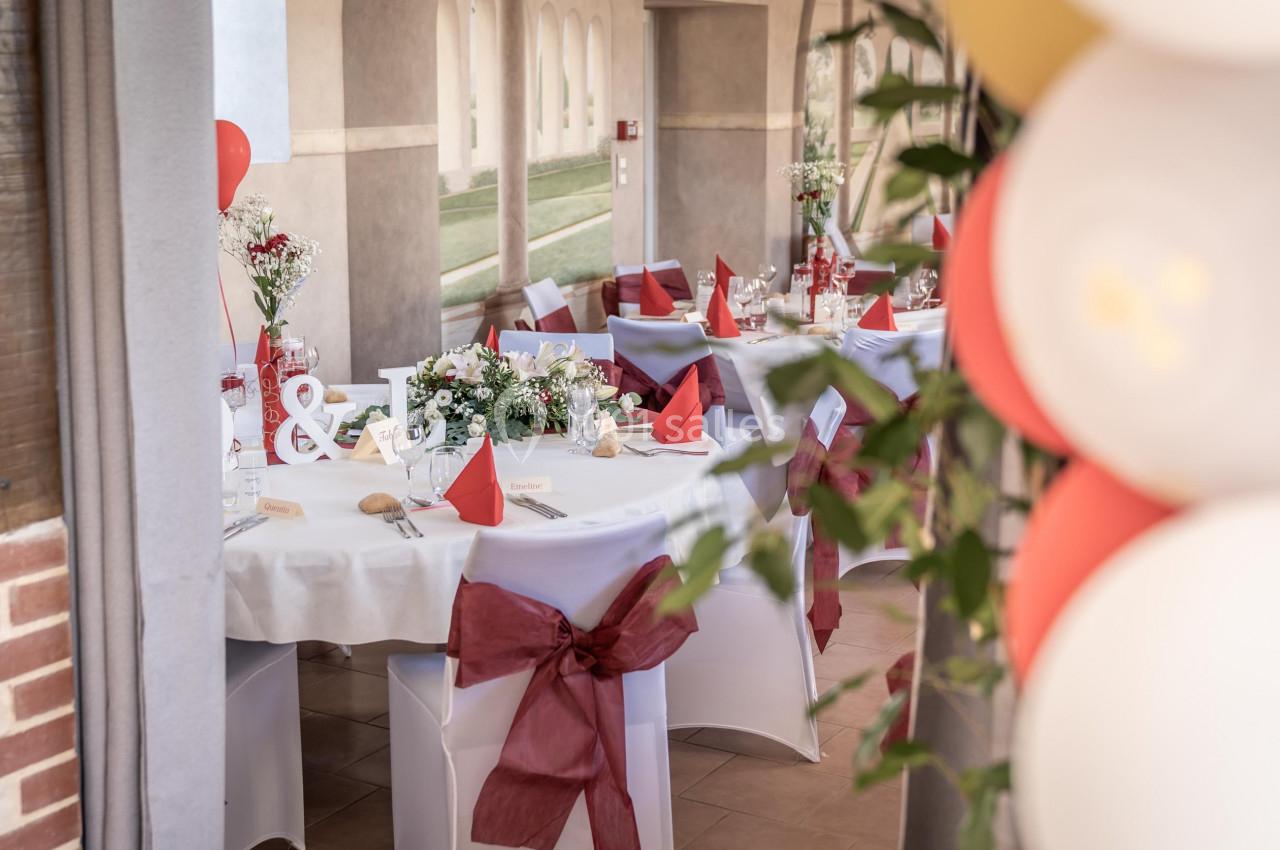 Salle décorée pour un événement avec tables dressées, nappes blanches, chaises ornées de nœuds rouges et ballons colorés.