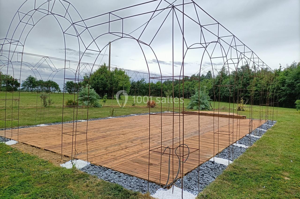 Structure métallique en arc installée sur une plateforme en bois, entourée de pelouse et d'arbres sous un ciel nuageux.