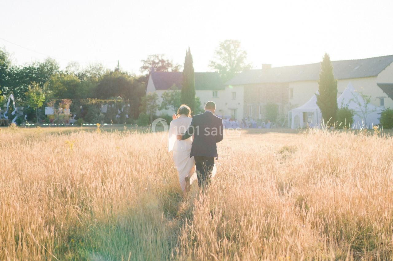 Un couple marche dans un champ doré au coucher du soleil, avec des maisons et des arbres en arrière-plan.