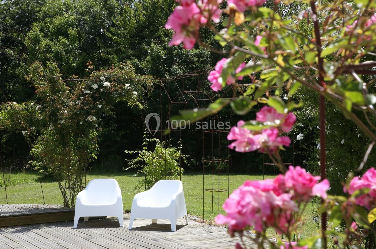 Deux fauteuils blancs sur une terrasse en bois, entourés de verdure et de fleurs roses au premier plan.