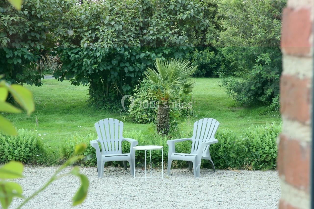Deux chaises blanches et une petite table sont disposées sur du gravier, face à un jardin verdoyant.