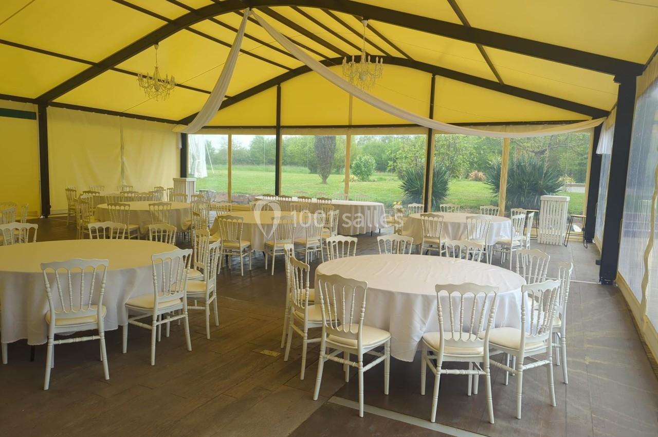 Salle de réception lumineuse avec tables rondes dressées, chaises blanches et vue sur un jardin verdoyant à travers des…