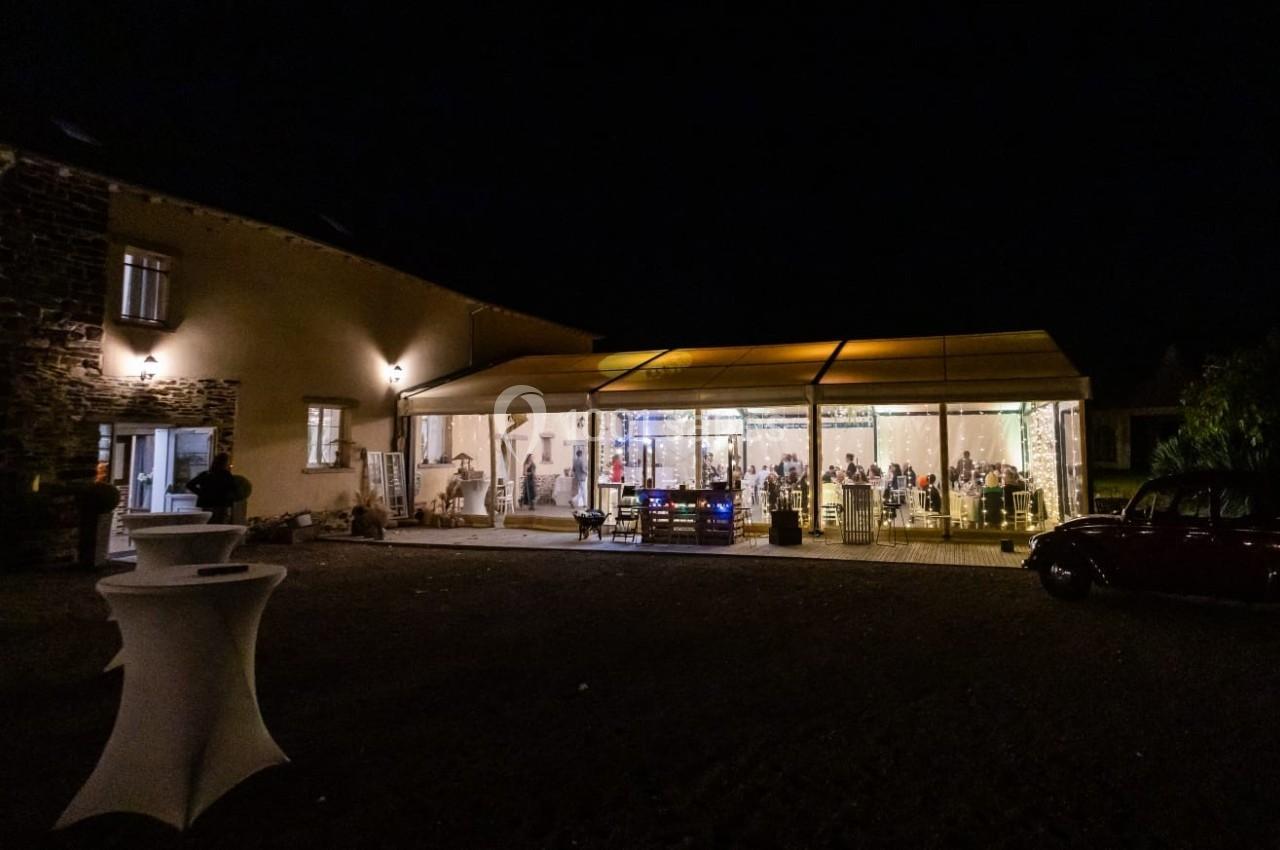 Salle de réception éclairée la nuit avec des invités visibles à l'intérieur, entourée d'un extérieur sombre.