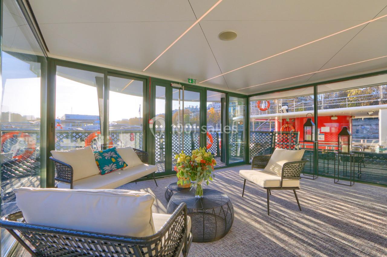 Salon lumineux avec mobilier en rotin, table basse et grandes baies vitrées donnant sur un bateau et un paysage urbain.