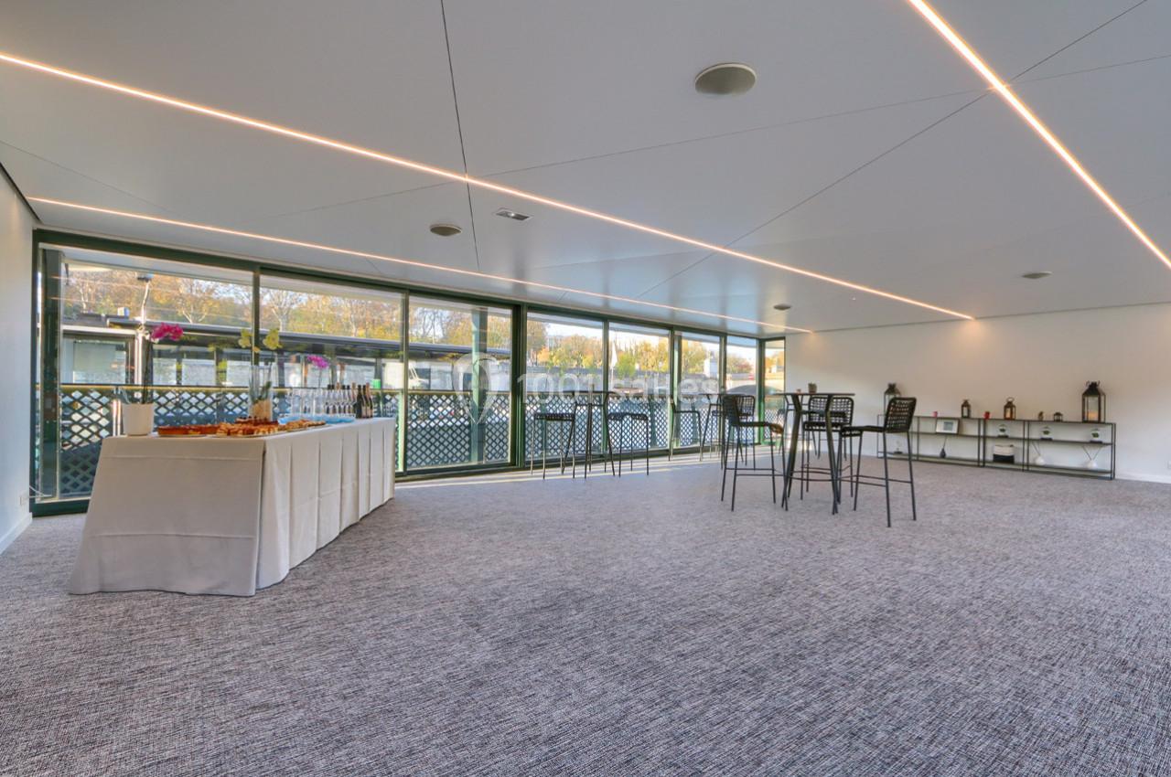 Salle lumineuse avec moquette grise, grandes baies vitrées, table de buffet et chaises hautes.