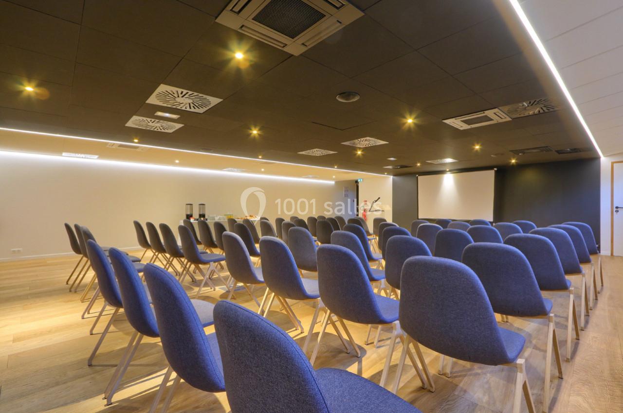 Salle de conférence moderne avec rangées de chaises bleues, écran de projection et éclairage au plafond.