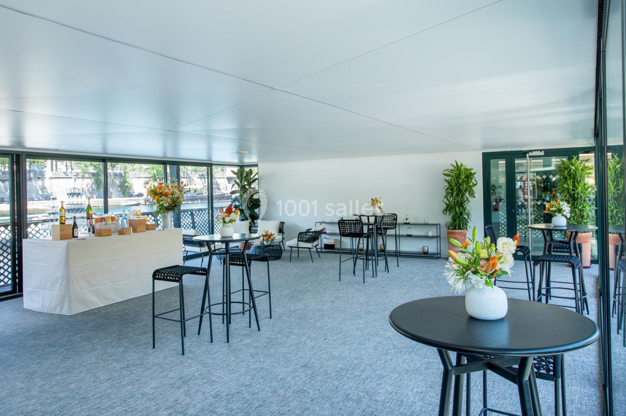 Salle lumineuse avec tables hautes, chaises noires, plantes décoratives et buffet dressé près d'une baie vitrée.