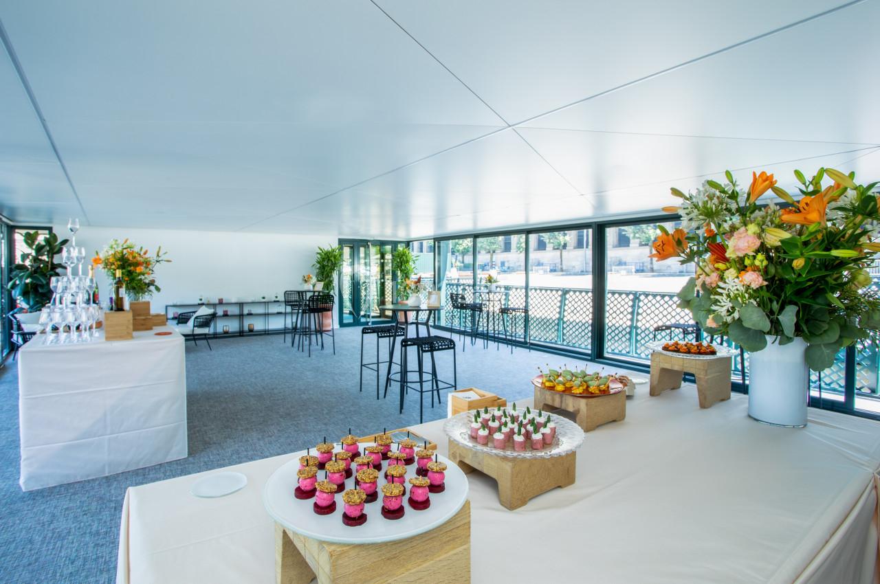 Salle lumineuse aménagée pour un événement, avec tables garnies de desserts et décorations florales.