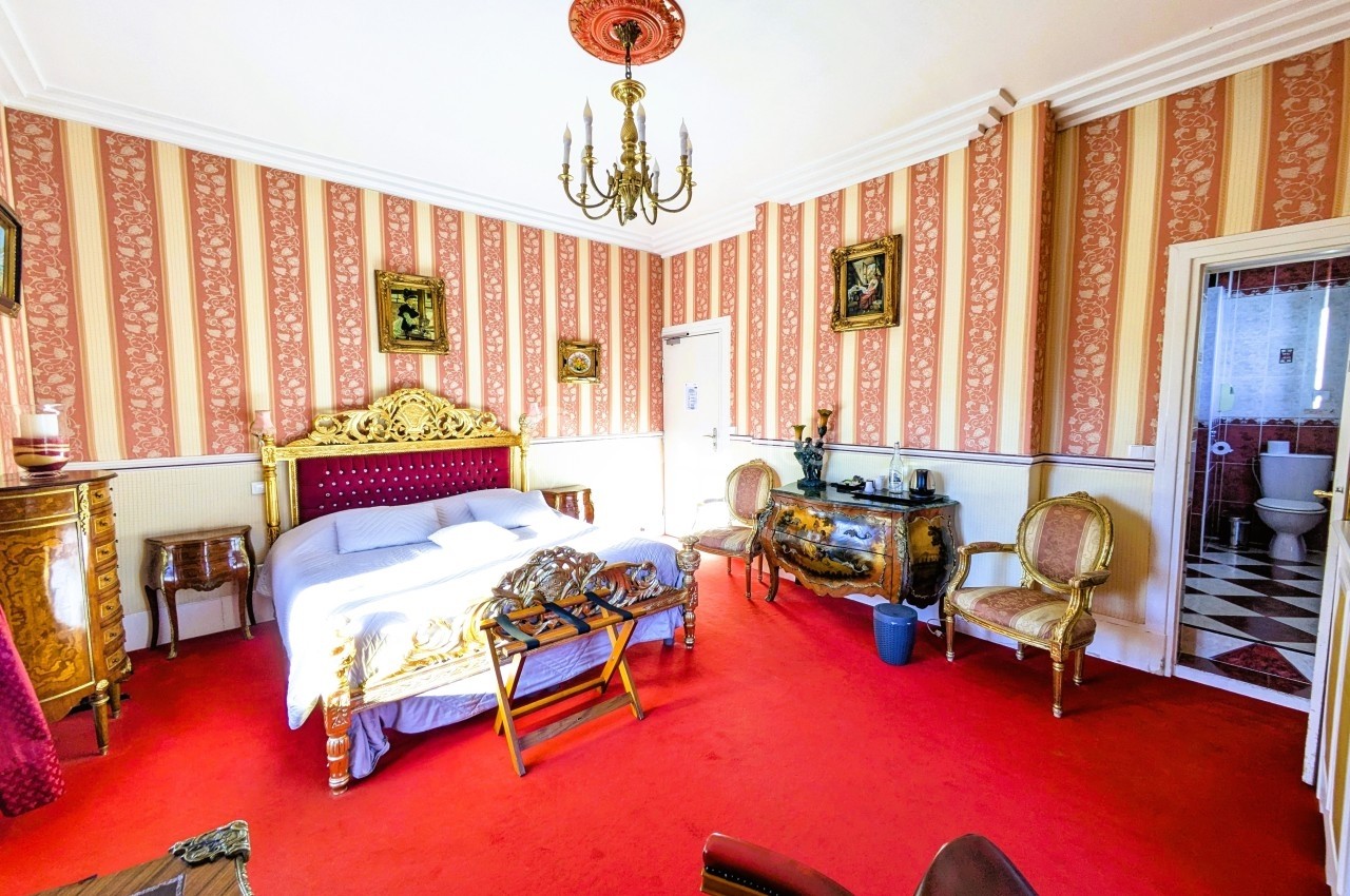 Chambre avec lit double, mobilier ancien, murs à rayures rouges et blanches, sol en moquette rouge, vue sur une salle de…