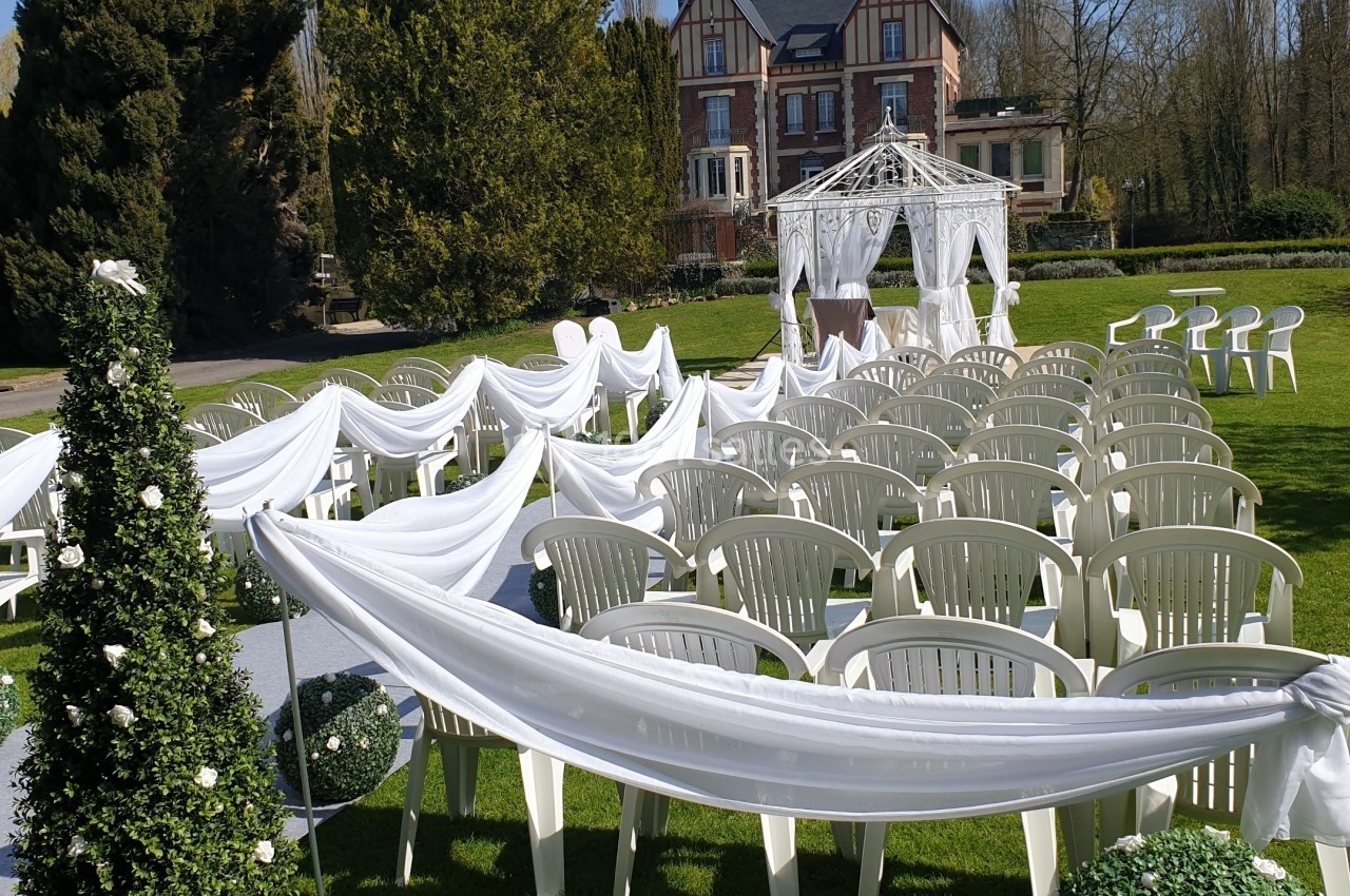 Chaises blanches alignées en extérieur avec des décorations en tissu blanc, face à une arche de mariage sur une pelouse.