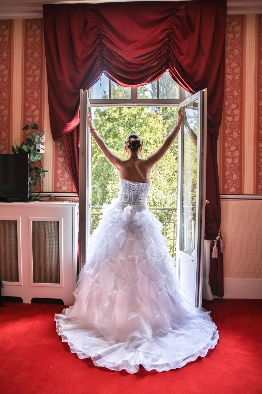 Une femme en robe de mariée blanche regarde par une porte-fenêtre ouverte donnant sur un paysage verdoyant.