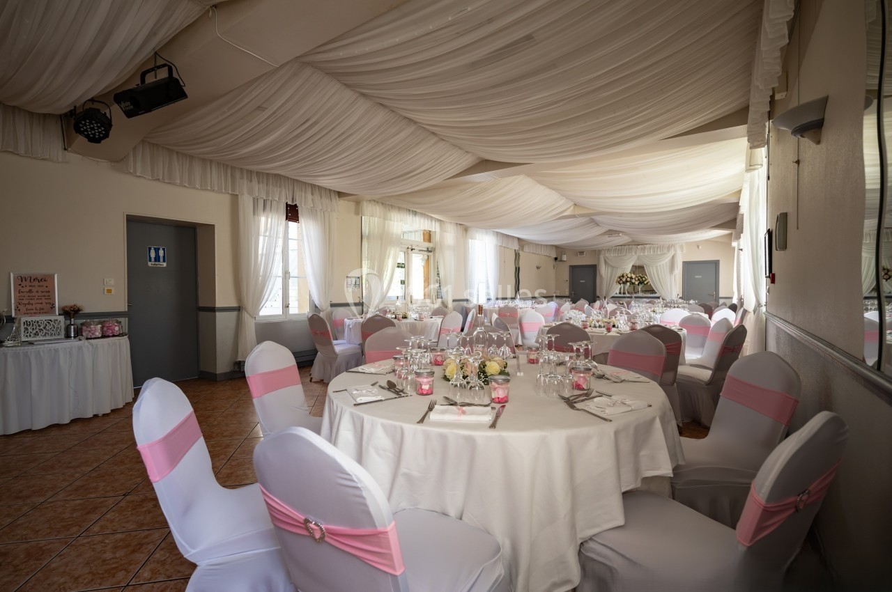 Salle de réception décorée pour un mariage, avec tables rondes dressées et chaises couvertes de housses blanches et roses.