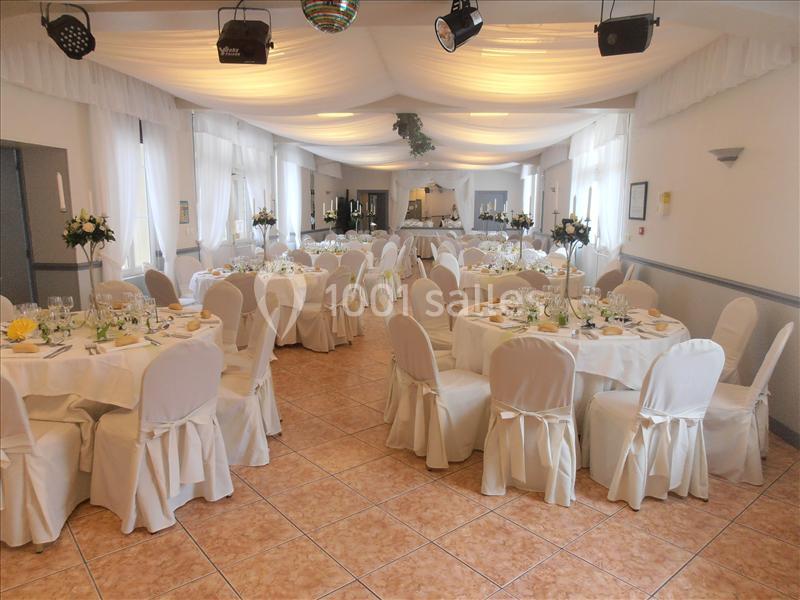 Salle de réception décorée avec des tables rondes dressées, nappes blanches et chaises couvertes, éclairage doux.