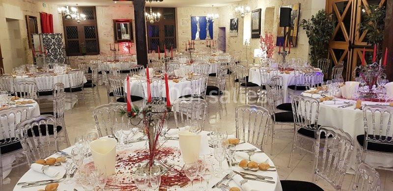 Salle de réception décorée avec des tables rondes dressées, chandeliers rouges et chaises transparentes.