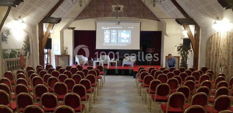 Salle de conférence avec des rangées de chaises rouges, une scène équipée de fauteuils et un écran de projection allumé.
