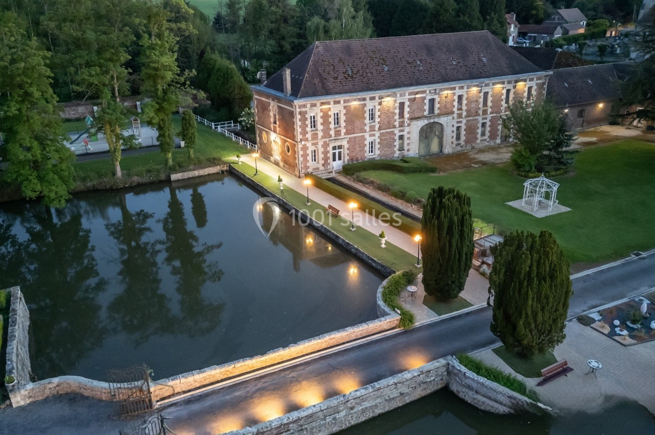 Vue aérienne d'un bâtiment en briques près d'un plan d'eau, entouré d'arbres et d'un jardin éclairé en soirée.