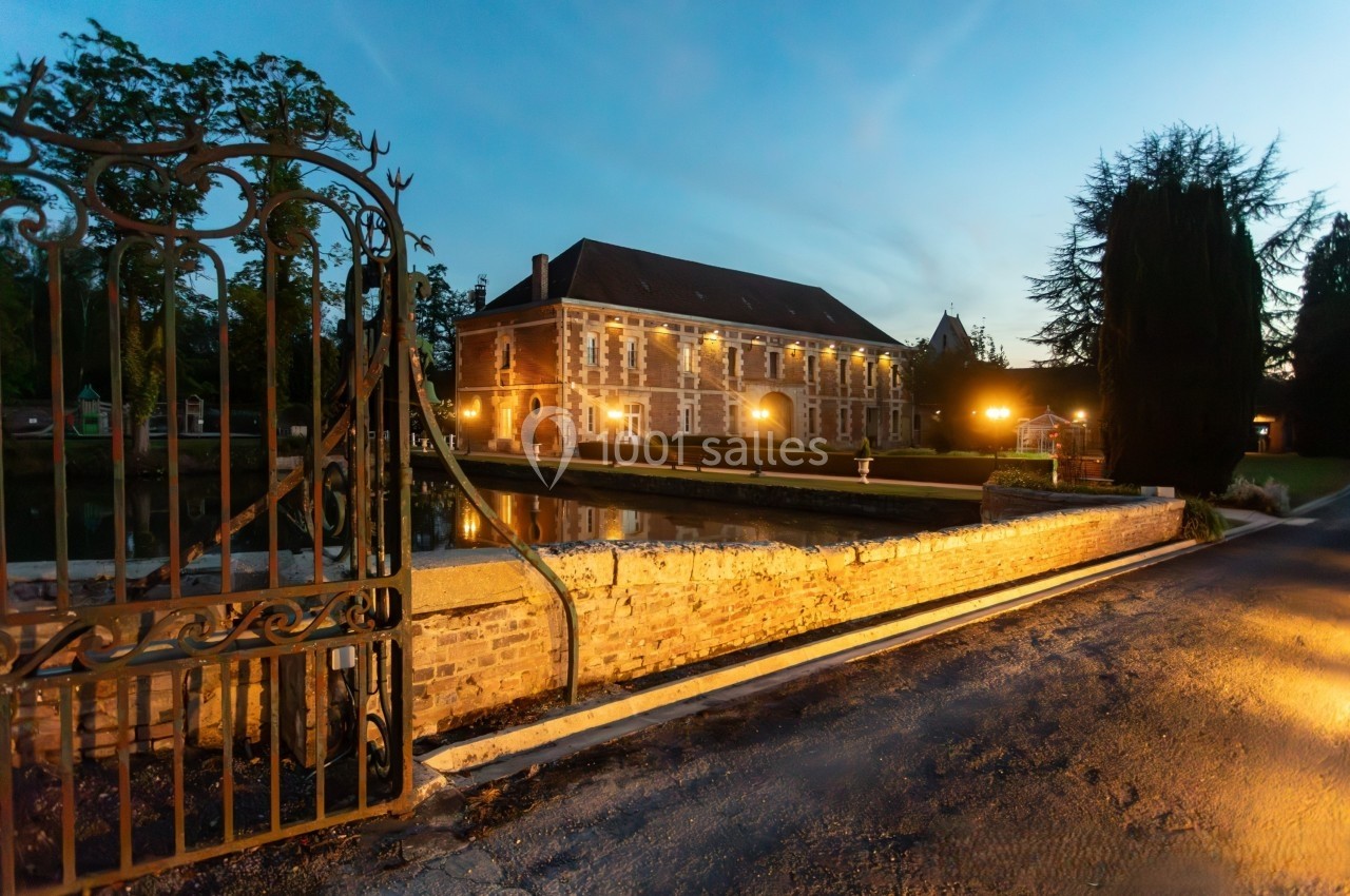 Manoir en briques éclairé au crépuscule, entouré d'un jardin, d'un étang et d'une grille en fer forgé.