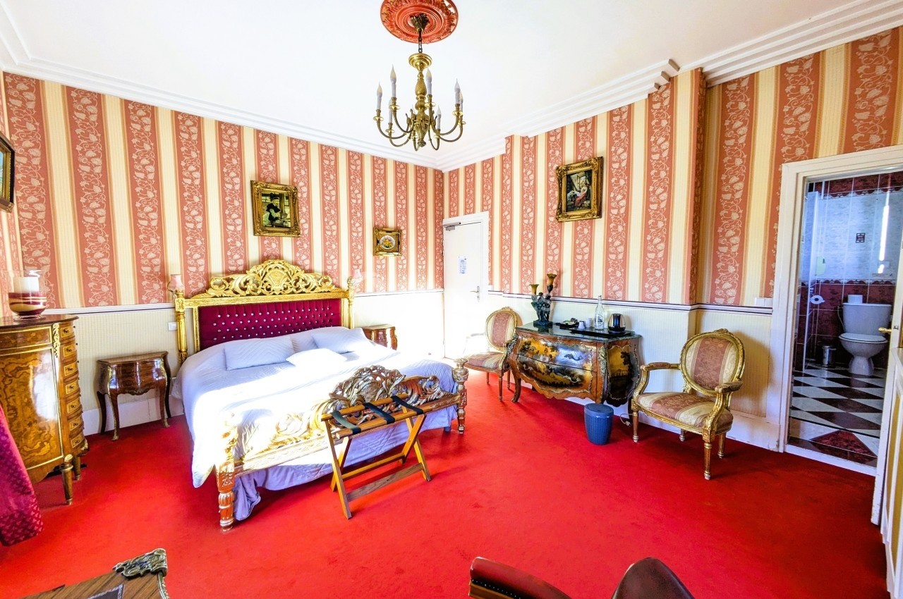 Chambre avec lit double, mobilier ancien, tapis rouge, murs rayés et vue sur une salle de bain avec carrelage noir et blanc.