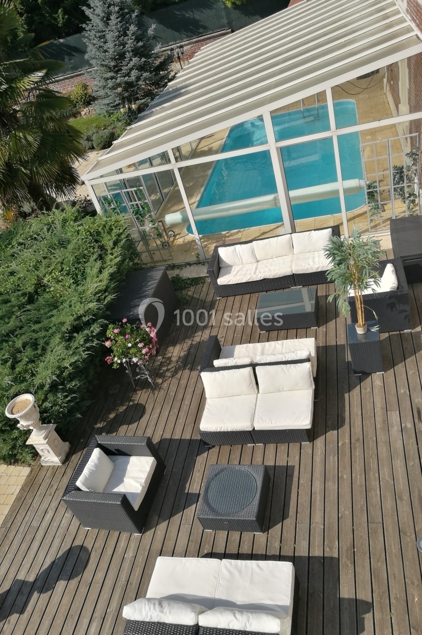 Terrasse en bois avec mobilier de jardin, vue sur une véranda abritant une piscine, entourée de végétation.