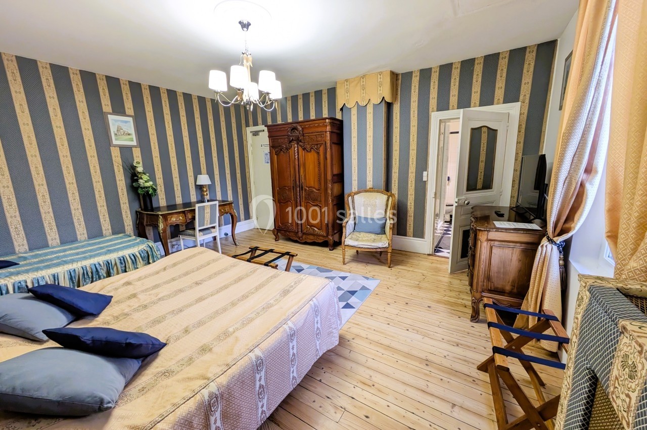 Chambre spacieuse avec lits, armoire en bois, bureau, fauteuil et décoration classique sur un parquet clair.