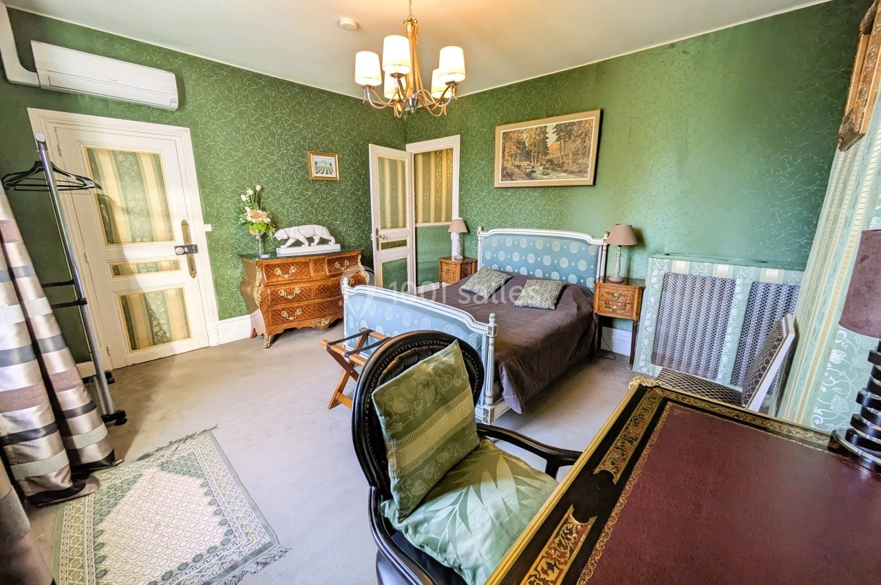 Chambre avec lit double, bureau, fauteuil, murs verts, mobilier classique et éclairage naturel provenant d'une fenêtre.