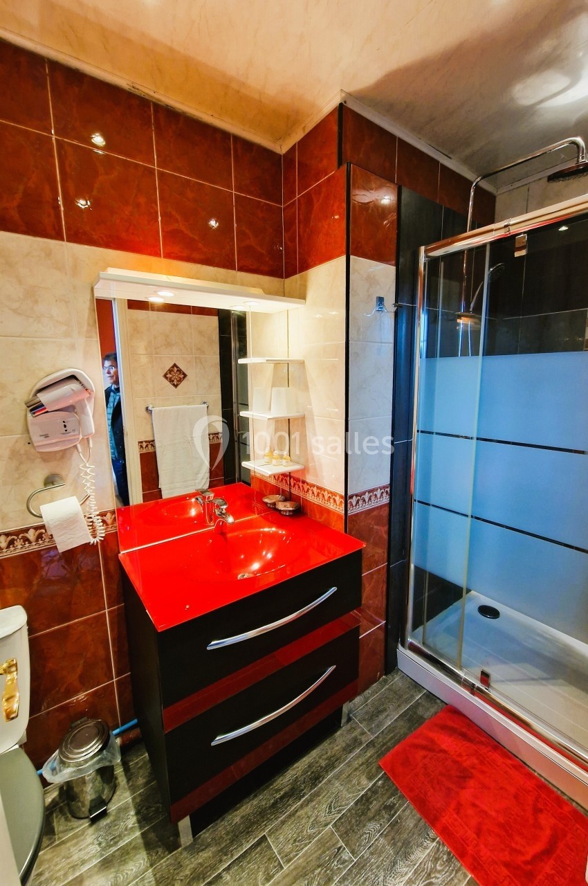 Salle de bain avec lavabo rouge, miroir éclairé, douche vitrée, carrelage beige et rouge, et sol effet bois.