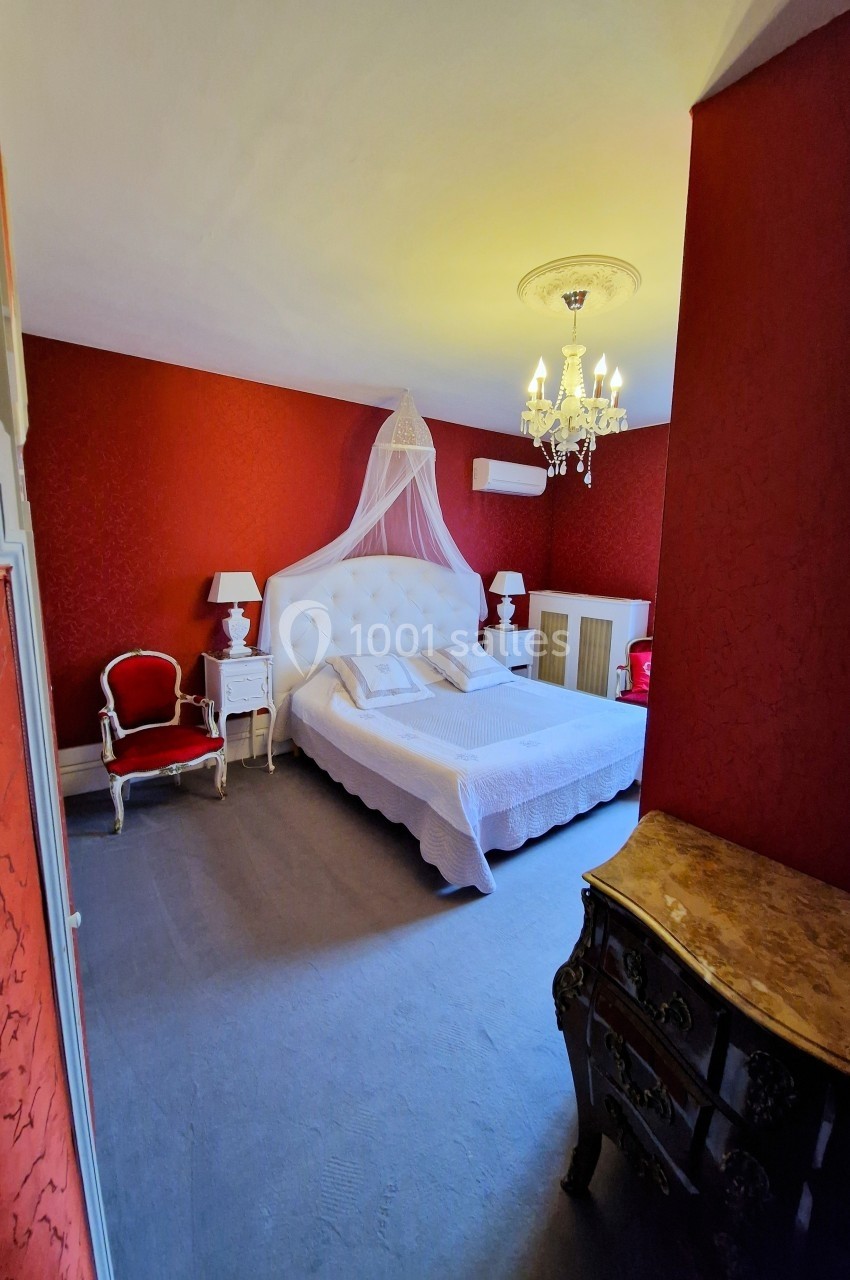 Chambre avec murs rouges, lit double à baldaquin blanc, mobilier classique et lustre au plafond.