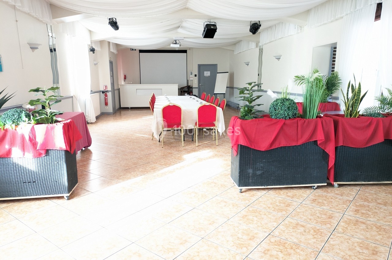 Salle lumineuse avec tables, chaises rouges et plantes décoratives, sous un plafond drapé de tissu blanc.