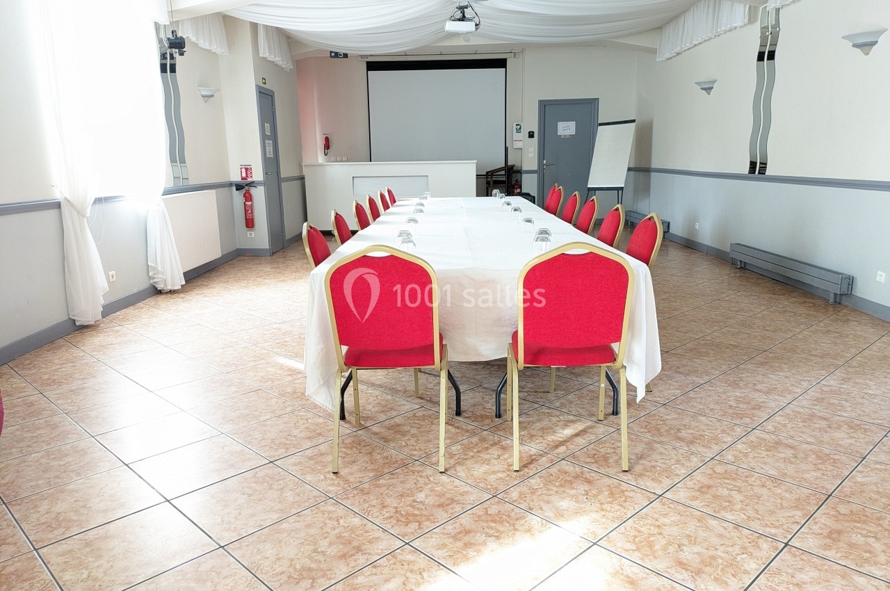Salle lumineuse avec une grande table rectangulaire couverte d'une nappe blanche et entourée de chaises rouges.