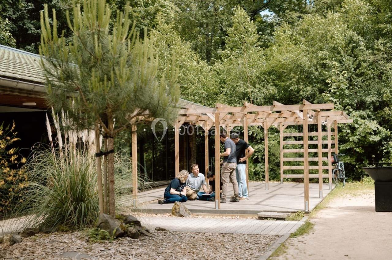 Un groupe de personnes discute sous une pergola en bois, entourée de végétation, près d'un bâtiment.