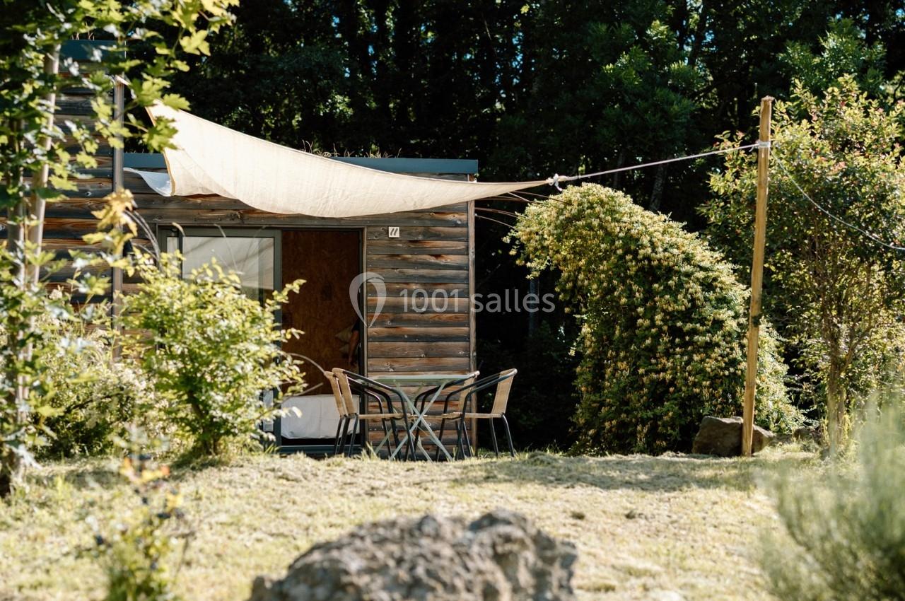 Table et chaises en extérieur devant une petite habitation en bois, entourée de végétation et ombragée par une toile tendue.
