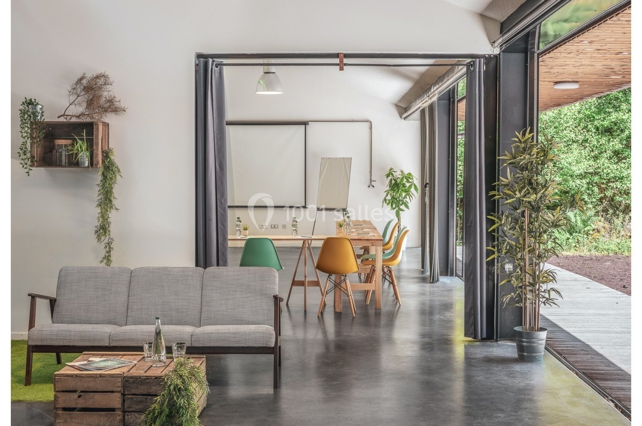 Salle lumineuse avec canapé, table en bois, chaises colorées, plantes, et grande ouverture sur un espace extérieur verdoyant.