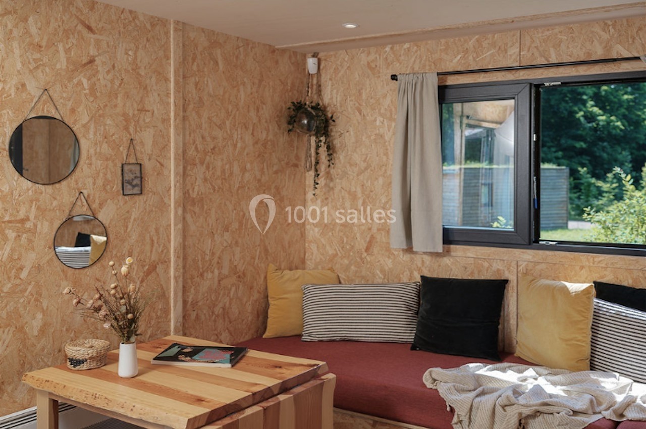 Salon lumineux avec murs en bois, canapé rouge, coussins colorés, table basse en bois et fenêtre donnant sur un jardin.