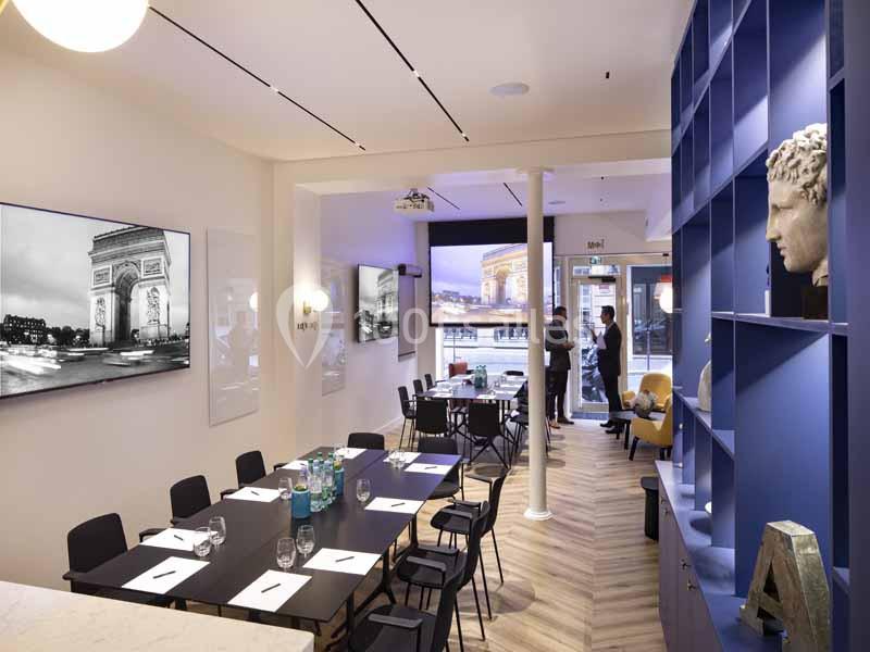 Salle de réunion moderne avec table, chaises, écrans muraux et étagères décorées, deux personnes discutant au fond.