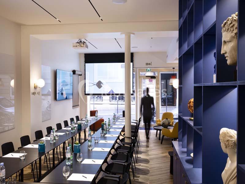 Salle de réunion lumineuse avec tables alignées, bouteilles d'eau, écran de projection et étagères décoratives.