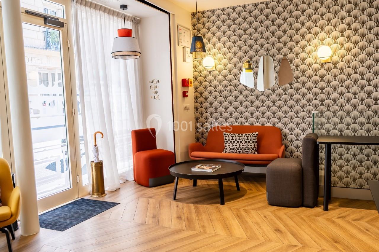 Salon moderne avec canapé orange, table basse ronde, chaises, lampes suspendues et papier peint à motifs géométriques.